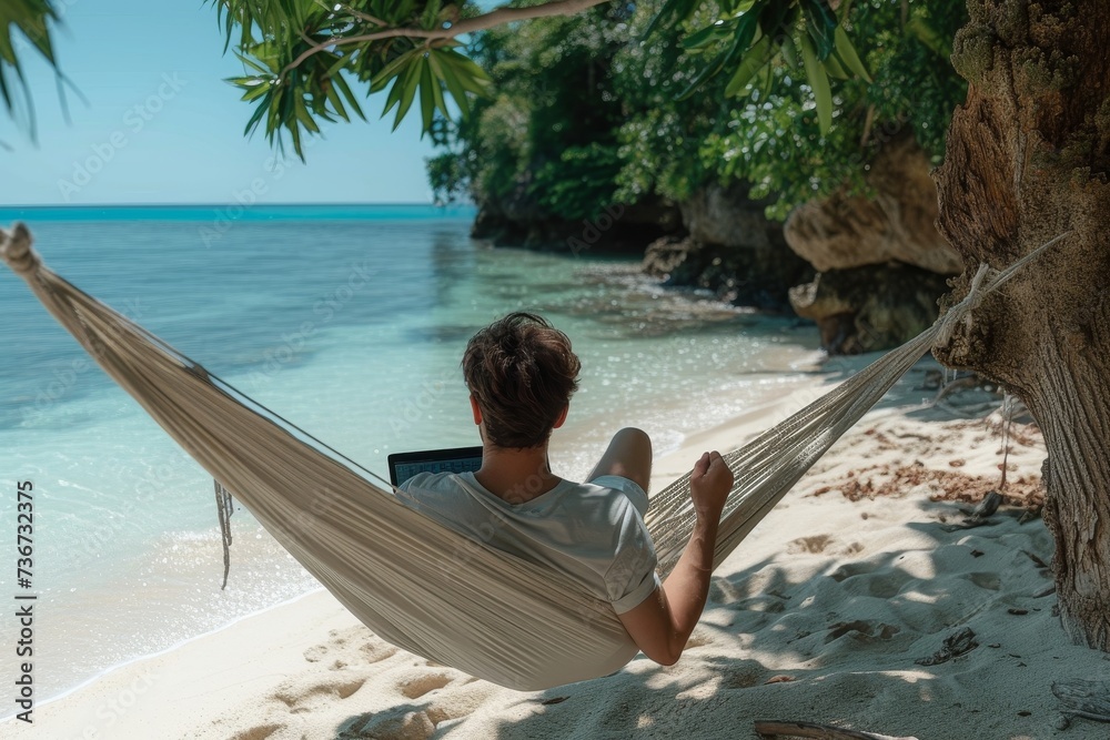 The back view of a man enjoying remote work from a hammock under the shade of palm trees with a sea view