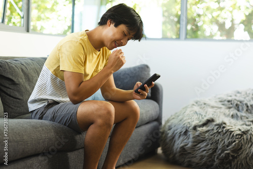Teenage Asian boy sits comfortably on a couch at home, smiling at his phone photo