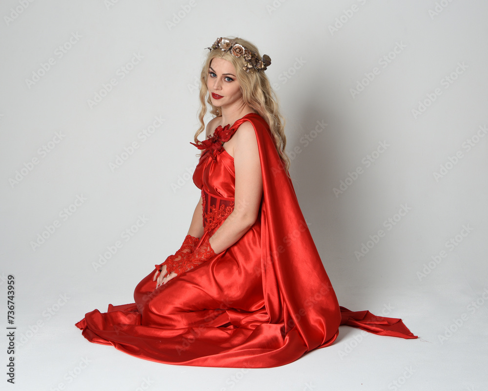 Full length portrait of beautiful blonde model dressed as ancient mythological fantasy goddess in flowing red silk toga gown, crown. Kneeling pose sitting on floor. isolated on white studio background Full length portrait of beautiful blonde model dressed as ancient mythological fantasy goddess in flowing red silk toga gown, crown. Kneeling pose sitting on floor. isolated on white studio background