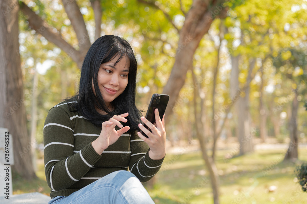 A positive young Asian woman using her smartphone while relaxing on a bench in a green park. A positive young Asian woman using her smartphone while relaxing on a bench in a green park.