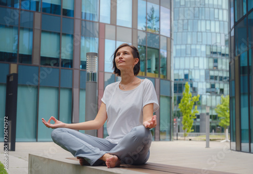 De-stress, unwind idea. peaceful female freelance employee practicing yoga exercises while sitting in bunch under glass skyscrapers after hard working day in office, holding hands in mudra