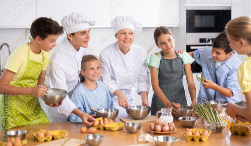 During lesson in cooking courses, female cook professional tells children about rules ticks and tricks for making fluffy pancakes, guy assistant helps hesitant child mix ingredients in bowl photo