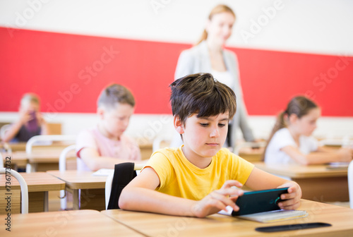 Focused diligent tween schoolboy sitting at lesson in classroom, using mobile phone