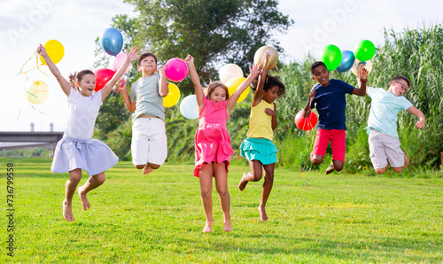 Group of happy kids jumping on grass with balloons in hands.