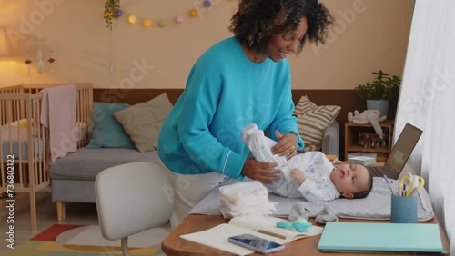 Medium shot of young African American woman taking off baby onesie while changing diaper to her infant son lying on changing table indoors photo