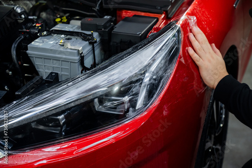 The master applies vinyl film to the headlight of a red car. Closeup view on worker detailer hand smoothing with a scraper protective film. 