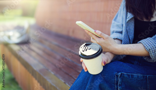 Young asian woman in casual clothes sitting on a bench using mobile phone enjoying and other hand holding coffee cup in her summer holiday
