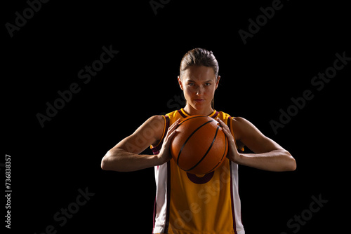 Young Caucasian female basketball player holds a basketball confidently on a black background