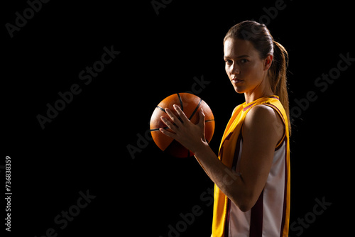 Young Caucasian female basketball player holds a basketball confidently on a black background, with 