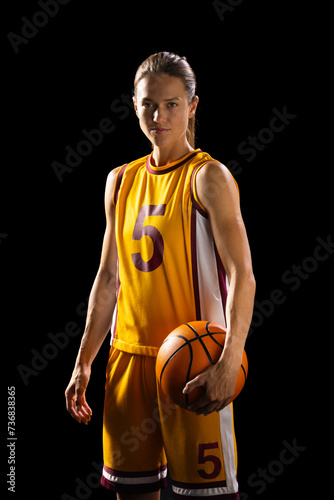 Athletic young Caucasian female basketball player poses confidently in basketball gear on a black ba