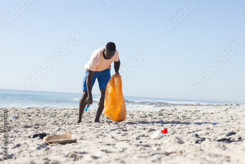 A young African American man cleans a beach, collecting trash, with copy space