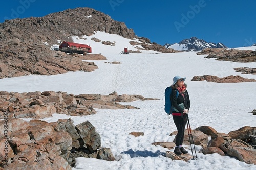 Middle-aged woman hiker relaxing and enjoying the view of the mountains covered with snow. Red hut on the background