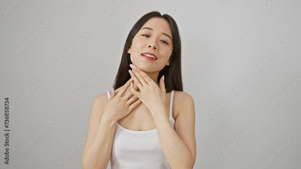 A young asian woman with a serene expression poses in a tank top against a white background, her hands gently touching her face and hair. A young asian woman with a serene expression poses in a tank top against a white background, her hands gently touching her face and hair.