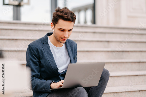 Concentrated young man wearing ear-pods is working on laptop while sitting outdoors. © Vulp