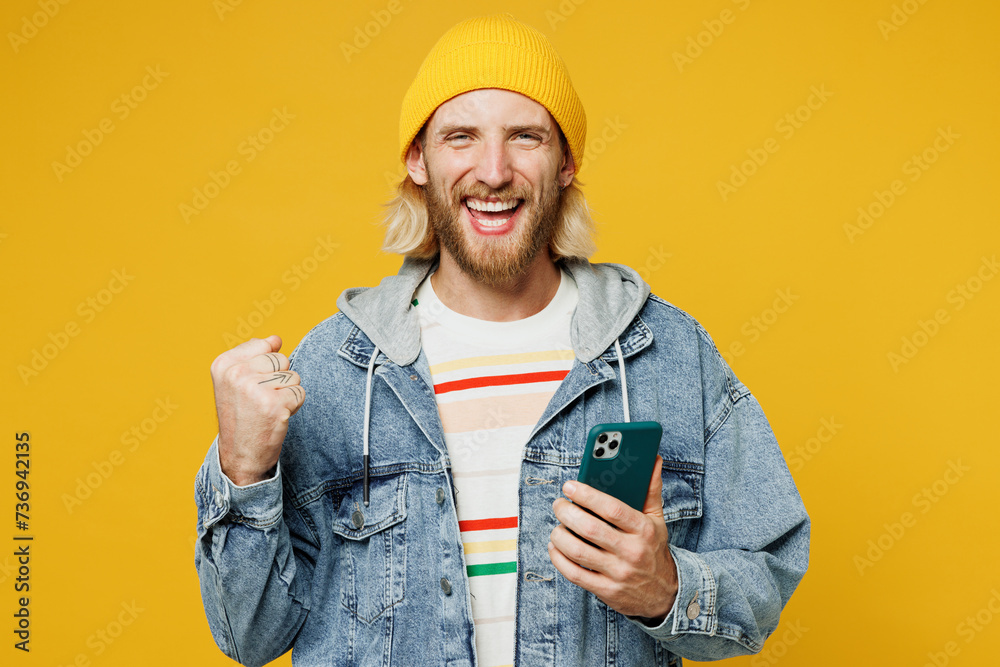 Young smiling happy blond man wears denim shirt hoody beanie hat casual clothes hold in hand use mobile cell phone show thumb up isolated on plain yellow background studio portrait. Lifestyle concept. Young smiling happy blond man wears denim shirt hoody beanie hat casual clothes hold in hand use mobile cell phone show thumb up isolated on plain yellow background studio portrait. Lifestyle concept.