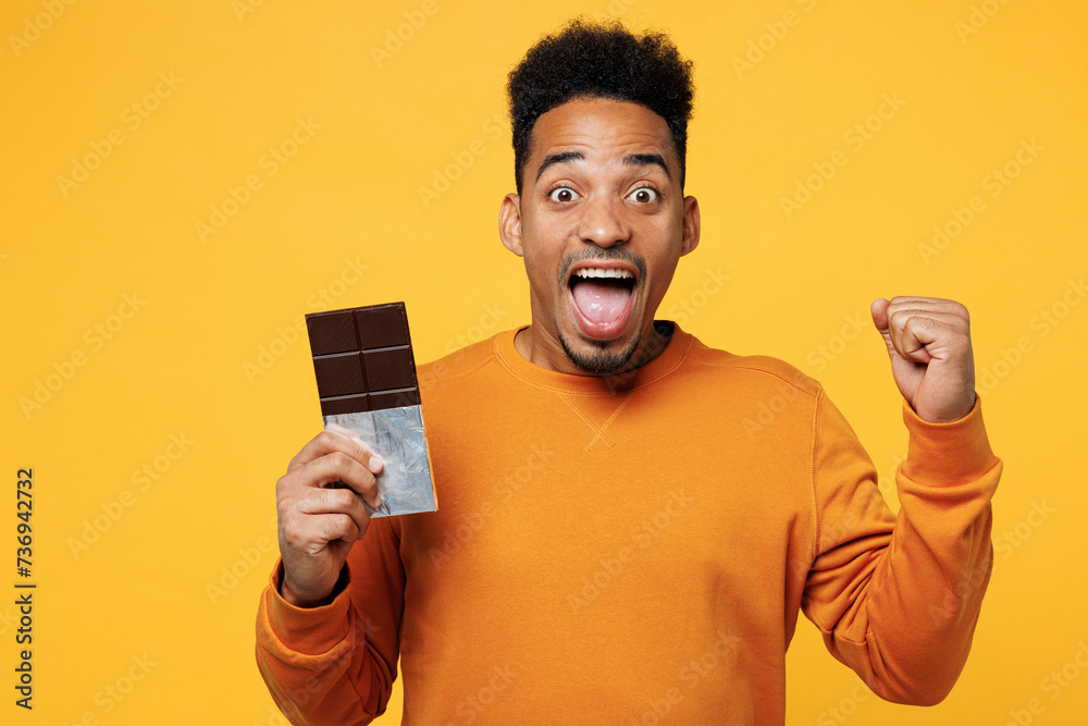Young surprised man wear orange sweatshirt casual clothes hold eat bar of chocolate do winner gesture isolated on plain yellow background. Proper nutrition healthy fast food unhealthy choice concept.