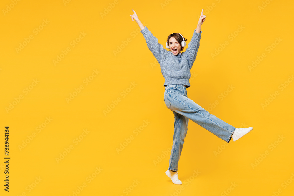 Full body side view young woman wears grey knitted sweater shirt casual clothes listen to music in headphones raise up hands leg isolated on plain yellow background studio portrait. Lifestyle concept.
