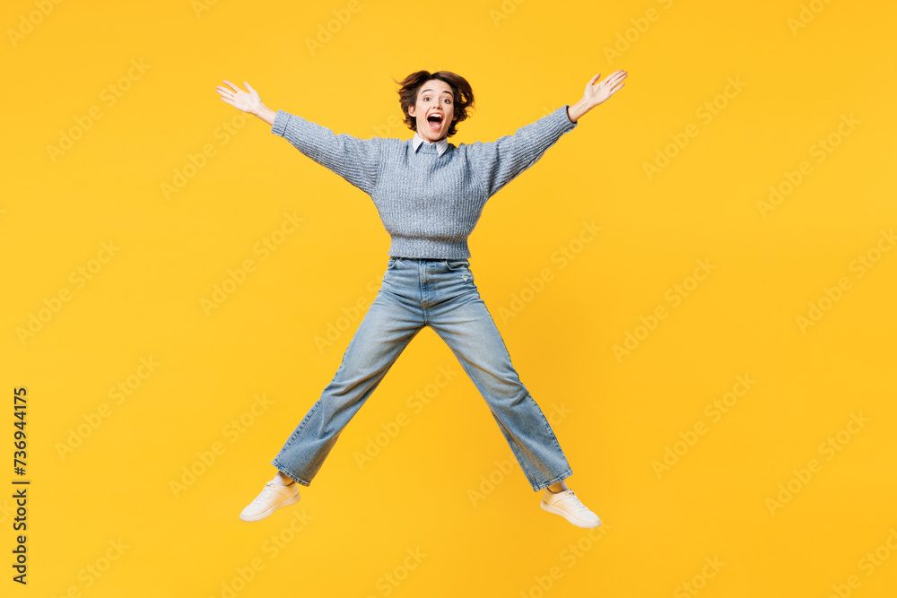 Full body astonished fun young woman she wears grey knitted sweater shirt casual clothes jump high with outstretched hands arms isolated on plain yellow background studio portrait. Lifestyle concept.