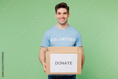 Young smiling happy man wears blue t-shirt white title volunteer hold cardboard donation box isolated on plain pastel green background. Voluntary free team work assistance help charity grace concept. photo