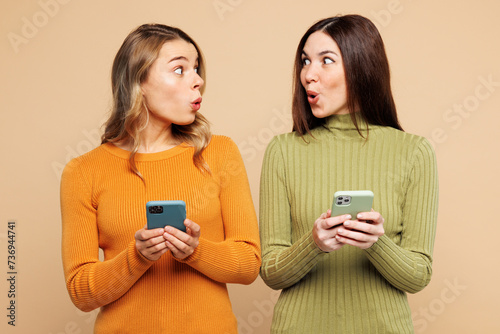 Young shocked amazed friends two women they wear orange green shirt casual clothes together hold in hand use mobile cell phone isolated on plain pastel light beige background studio Lifestyle concept photo