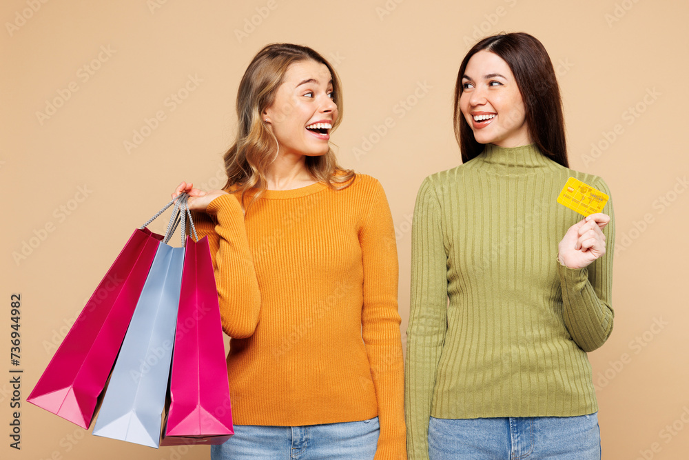 Young fun friends two women wear orange green shirt casual clothes hold in hand credit bank card shopping open paper package bags isolated on plain beige background. Black Friday sale buy day concept. Young fun friends two women wear orange green shirt casual clothes hold in hand credit bank card shopping open paper package bags isolated on plain beige background. Black Friday sale buy day concept.
