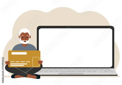A man sits cross-legged and holds a large plastic card. Next to the man is a large laptop with space for text.