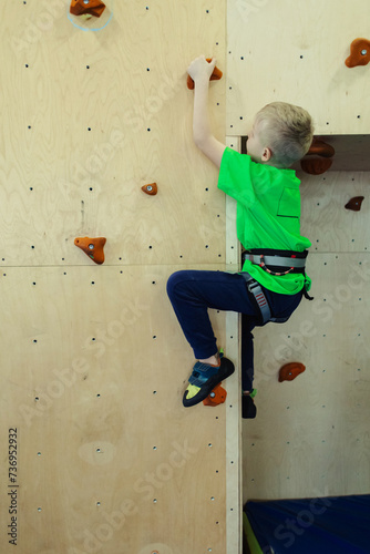 Rock climbing for children. Blond boy in a green shirt is climbing climbing wall. Bouldering class. Artificial track for safe activities and competitions. Mountaineering, safety system, special shoes