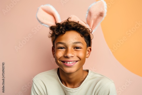 
Photography an indigenous boy, aged 15, smiling warmly with bunny ears on his head, against a gentle pastel peach backdrop, ideal for Easter-themed advertisements or social media posts photo