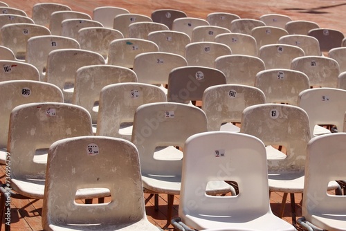 On display are rows of empty, simple, old, white-grey stacking chairs set up one behind the other for an outdoor event. The chairs stand on a dark wooden floor.