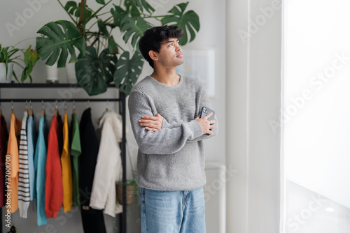 A contemplative young man stands by the window with his arms crossed, looking away thoughtfully. The indoor setting and casual clothing give a relaxed ambiance. photo