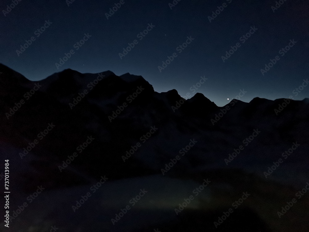 A breathtaking sight of towering mountains, illuminated by the soft glow of the moon in the distance at night.