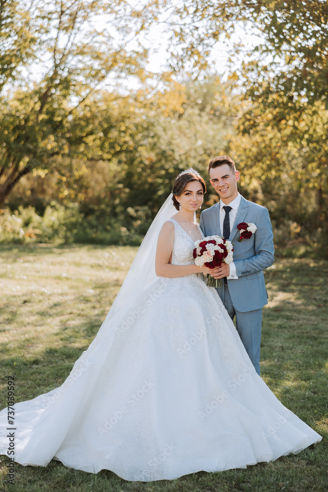 Stylish, young groom and beautiful bride in a long white dress and a long veil with a bouquet in their hands, hugging in the park in the autumn nature. Wedding portrait of newlyweds. Stylish, young groom and beautiful bride in a long white dress and a long veil with a bouquet in their hands, hugging in the park in the autumn nature. Wedding portrait of newlyweds.