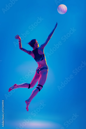 Dynamic portrait woman, sportsman, volleyball player training to hits ball in mid-air against gradient blue background in pink neon light. Concept of sport games, movement, active, healthy lifestyle. © Lustre
