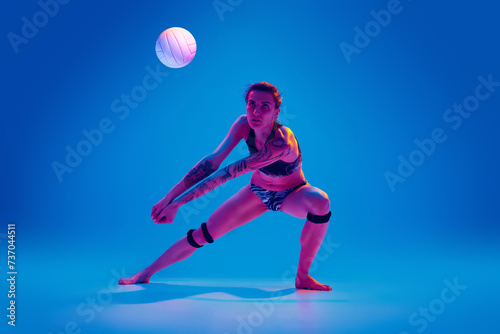 Young woman, volleyball player in motion, training, playing against gradient blue background in pink neon light. Concept of sport, movement, active and healthy lifestyle, power and strength.