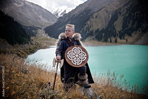 A young boy in the viking cosplay costume holding a wooden shield and a axe and standing next to the beautiful alpine lake with Mountains view