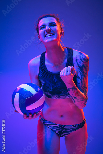 Female beach volleyball player clenches his hand into fist, rejoices at goal scored, holding ball against blue-purple background in neon light. Concept of sport, movement, victory, win, championship.