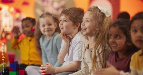 Group of Happy Excited Kids Sitting on a Floor and Watching Cartoons on a TV in a Modern Daycare Center. Diverse Children Having Fun, Learning Through Video Lessons, Games, Music, Art  photo
