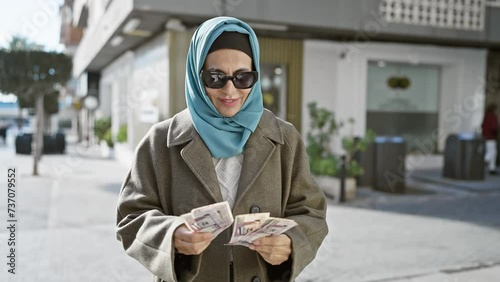 Mature woman with headscarf counting saudi riyal banknotes on urban street. photo