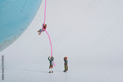 Photography of miniature people and toy figures, A mountaineer climbs up a globe on a rope and is observed by a team  photo