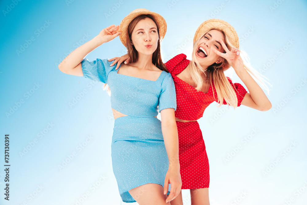 Two young beautiful smiling hipster woman in trendy summer same clothes. Carefree women in street in hats. Positive models at sunset. Cheerful and happy. Bottom view. They look at camera from above