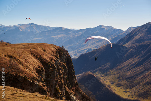Smoothly sailing in the clear sky, paragliders drift near brown mountain peaks.