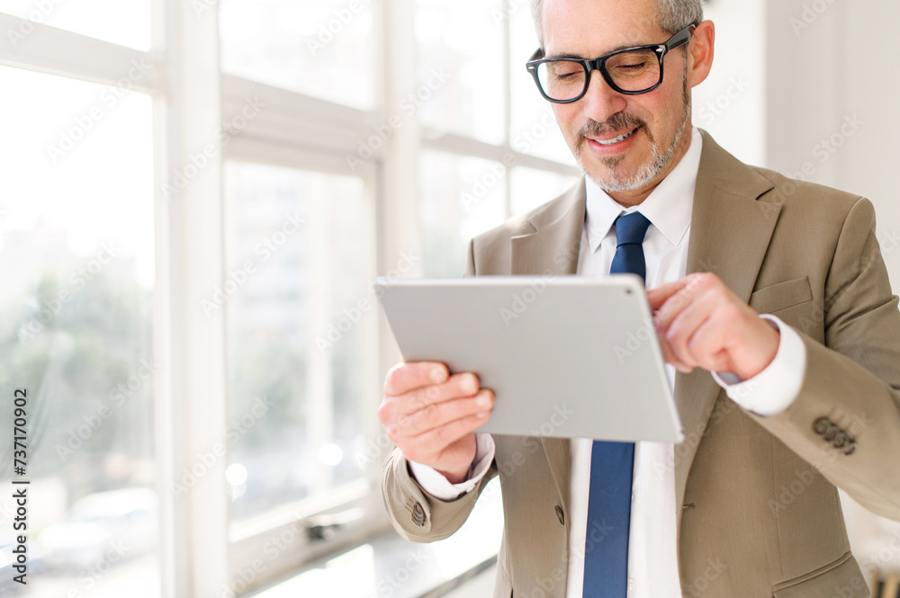 A senior businessman in a beige suit stands by a bright window, smiling as he engages with a tablet. This image captures the concept of modern technology in the hands of experienced professionals. A senior businessman in a beige suit stands by a bright window, smiling as he engages with a tablet. This image captures the concept of modern technology in the hands of experienced professionals.