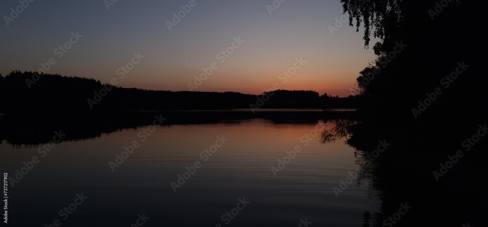 A freshwater reservoir at sunset is a captivating scene where the sun descends below the horizon, casting warm hues across the sky and reflecting on the tranquil water surface. The merging colors crea