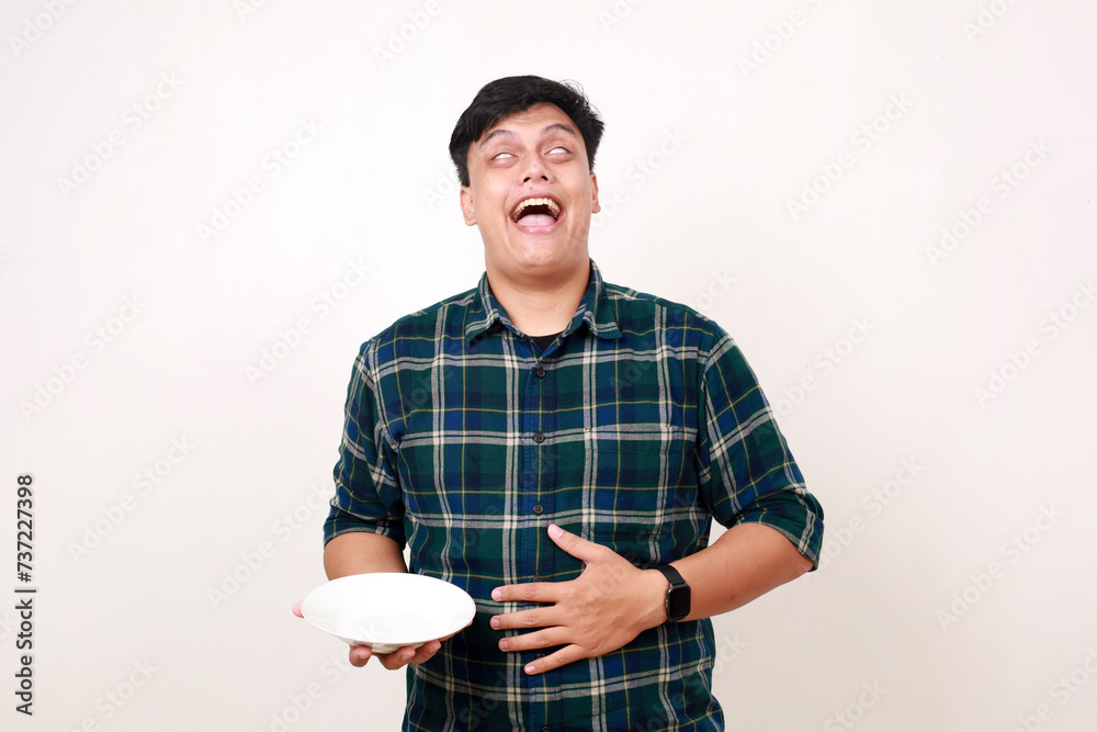 Satisfied Young asian man feels full, touching his stomach while holding an empty plate Satisfied Young asian man feels full, touching his stomach while holding an empty plate