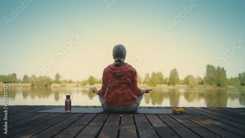 Relaxed grey-haired woman sitting in lotus pose on wooden pier, practicing yoga