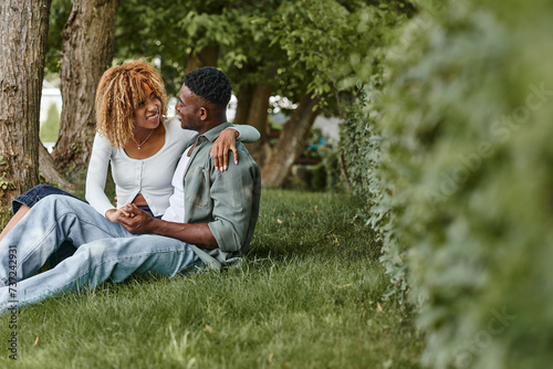 happy african american woman in casual wear hugging boyfriend while sitting together on grass