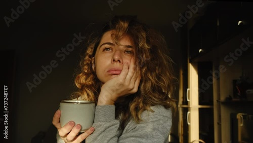 Creative crisis. Morning portrait of young woman. Lady holds mug of morning coffee, sighs heavily while sitting in sunny room at sunrise.