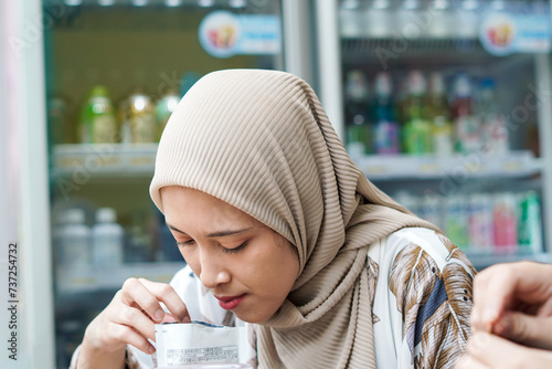 Indonesian Hijab Woman Pouring Packaged Beverage into Cup with Ice Cubes at Cafe or Minimarket photo