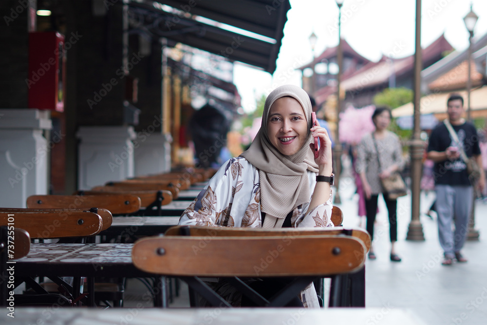 Fototapeta premium Indonesian Woman wearing Hijab Engaging in Digital Conversations at Cafe Bench