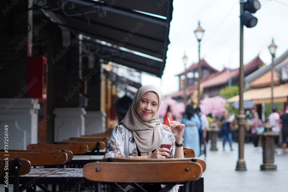 Indonesian Hijabi Woman Smiling with Refreshment at Cafe Bench. Joyful Moments at the cafe concept. Indonesian Hijabi Woman Smiling with Refreshment at Cafe Bench. Joyful Moments at the cafe concept.